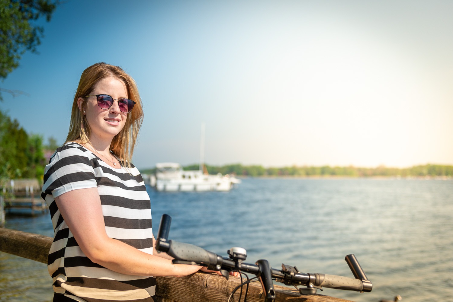 Eine junge Frau mit Sonnenbrille und gestreiftem T-Shirt steht mit einem Fahrrad auf einem Steg an der Havel in Werder (Havel) und schaut aufs Wasser.